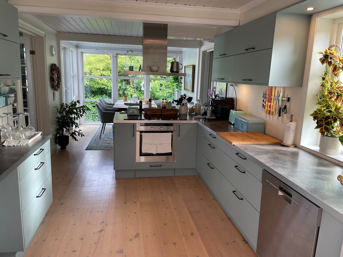 A modern kitchen features light blue cabinetry complemented by a wood countertop. Natural light streams through large windows, illuminating the space. A stainless steel oven and a variety of utensils are organized neatly, while an inviting dining area can be seen in the background.