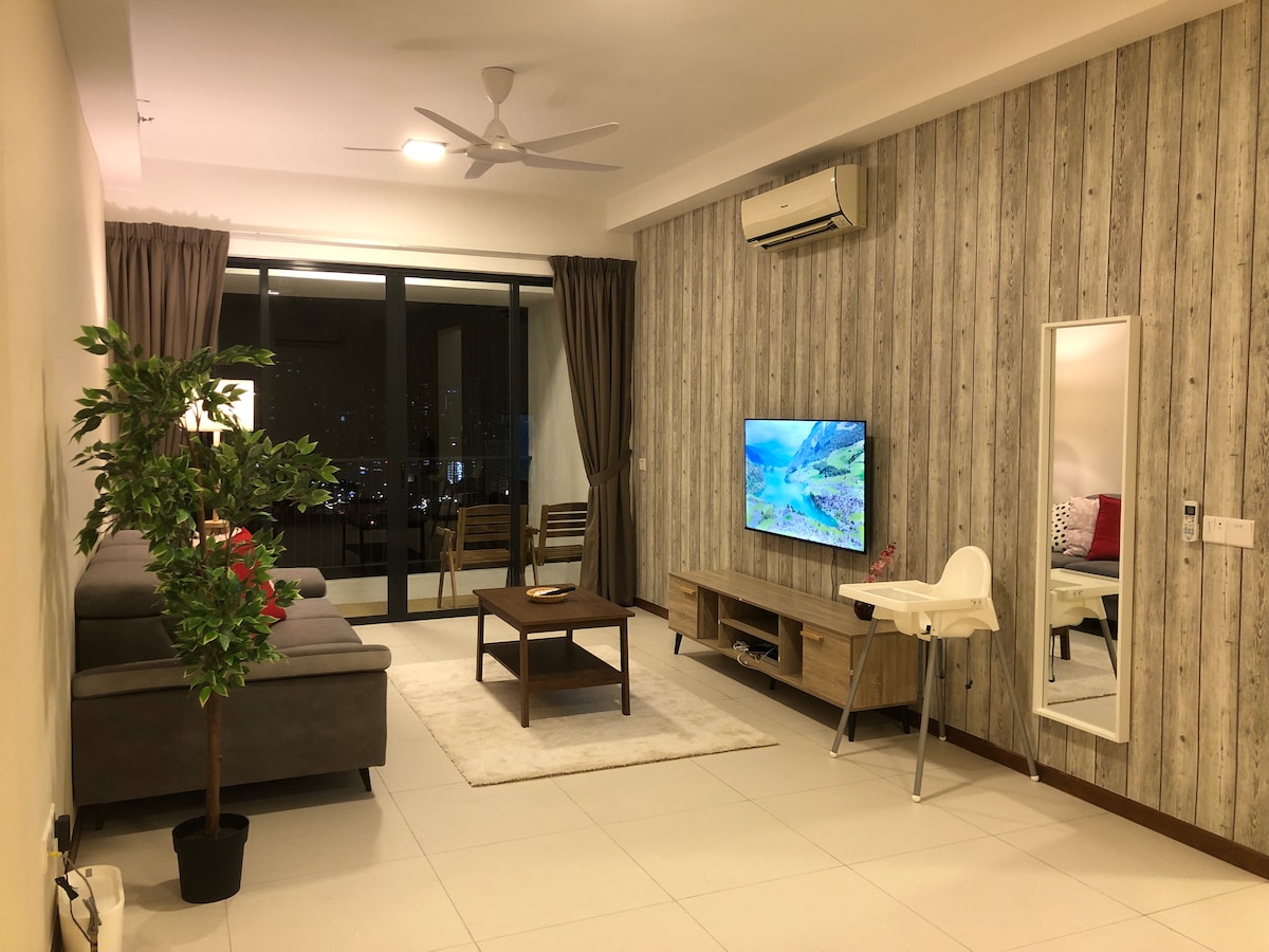 A cozy living area is presented with a gray sofa facing a wall-mounted television. A coffee table sits on a light rug, and a small dining area with wooden chairs is visible near a glass balcony door. Natural light is welcomed by wide windows, enhancing the space.