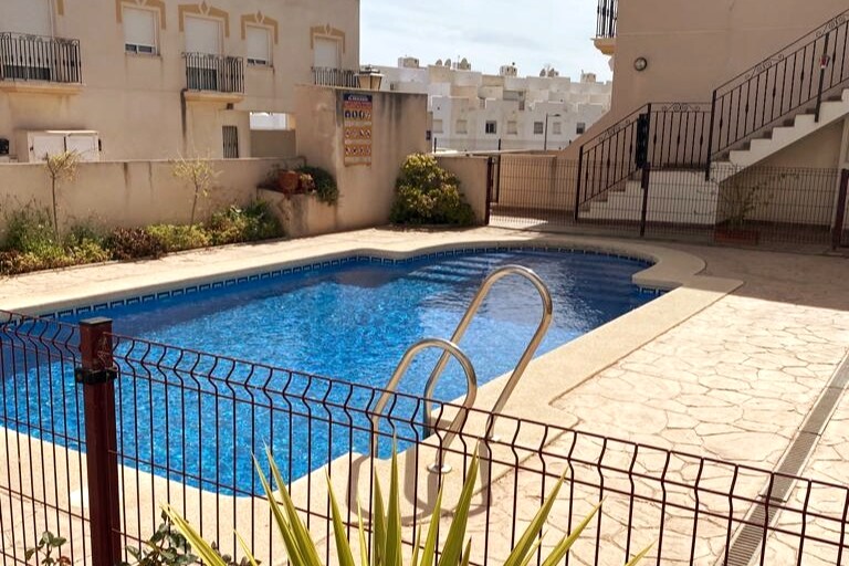 A swimming pool is framed by a decorative metal fence, with steps leading up to nearby apartments. Lush greenery and small shrubs are visible around the pool area. The pool's clear water reflects sunlight, creating a serene setting for relaxation.