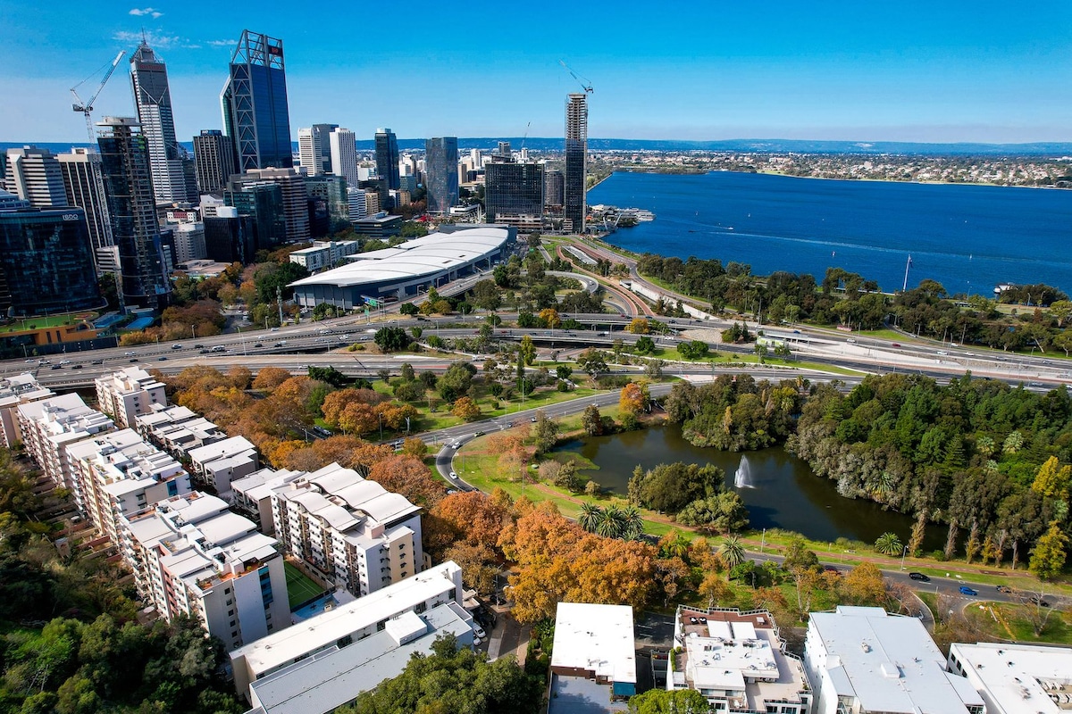An aerial view captures the vibrant landscape of Perth, featuring the sparkling waters of the river and the city skyline in the background. Green parks and autumn-colored trees contrast with the urban structures, while roads and pathways connect the various areas, showcasing the city’s layout and accessibility.
