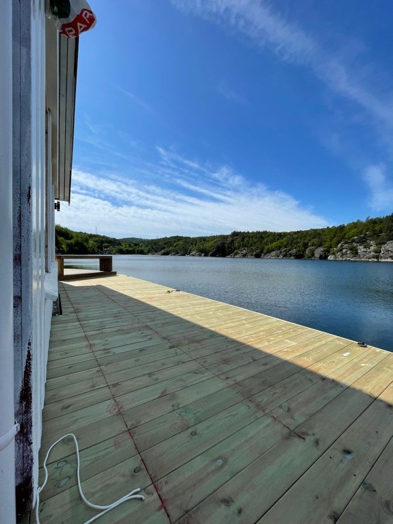 A wooden deck extends along the water's edge, providing a view of the calm lake surrounded by greenery and rocky hills. The clear sky above is brightened by soft clouds, creating a serene atmosphere ideal for relaxation.