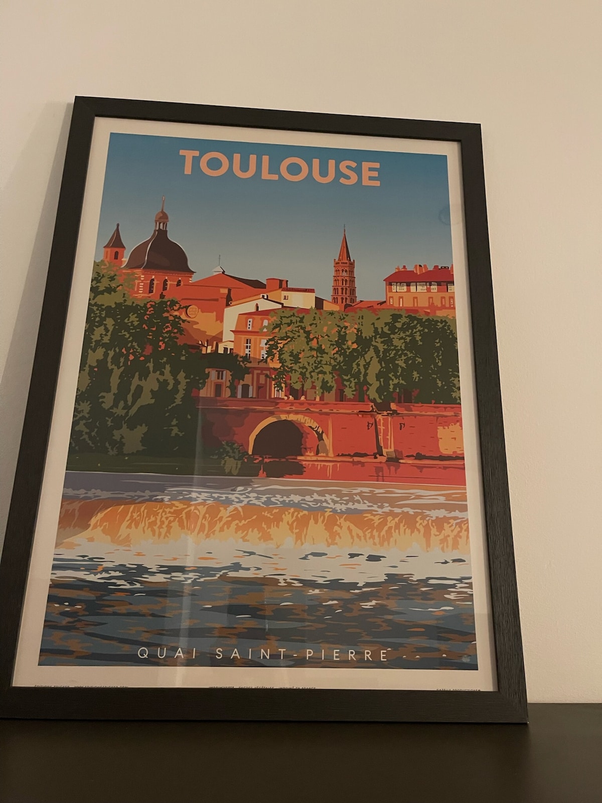 A framed art poster of Toulouse decorates the wall. The illustration depicts the cityscape with notable architectural features such as domed buildings and a spire, set against a backdrop of a flowing river. The text 'TOULOUSE' is prominently displayed at the top.