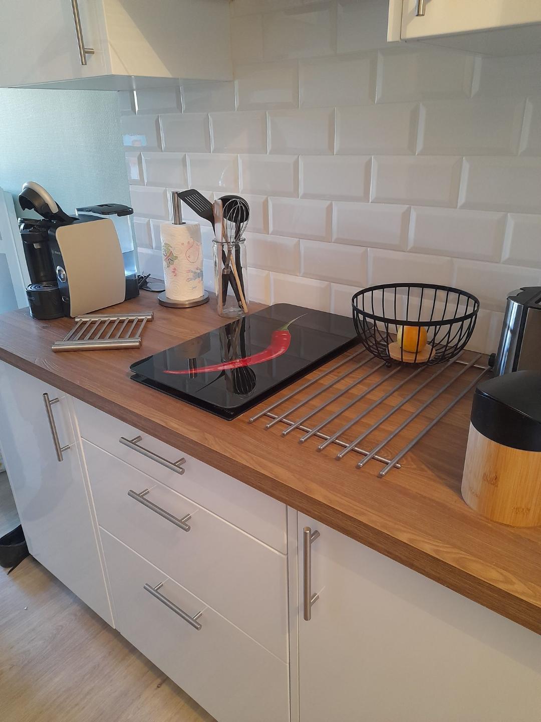 A modern kitchen counter is displayed, featuring a vitroceramic cooking plate, coffee machine, and various utensils. A wooden cutting board and a fruit bowl with an orange are visible. Storage cabinets with sleek handles complement the light-colored backsplash tiles.