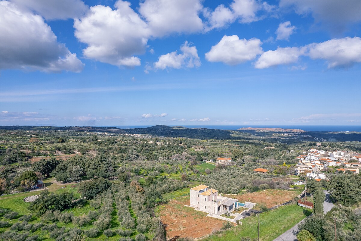 A panoramic view captures the tranquil landscape surrounding Mia Casa Villa, featuring lush greenery and distant hills under a blue sky with scattered clouds. The villa is positioned amidst open fields, with the coastline visible in the background, offering a glimpse into the peaceful environment of Rethymno.