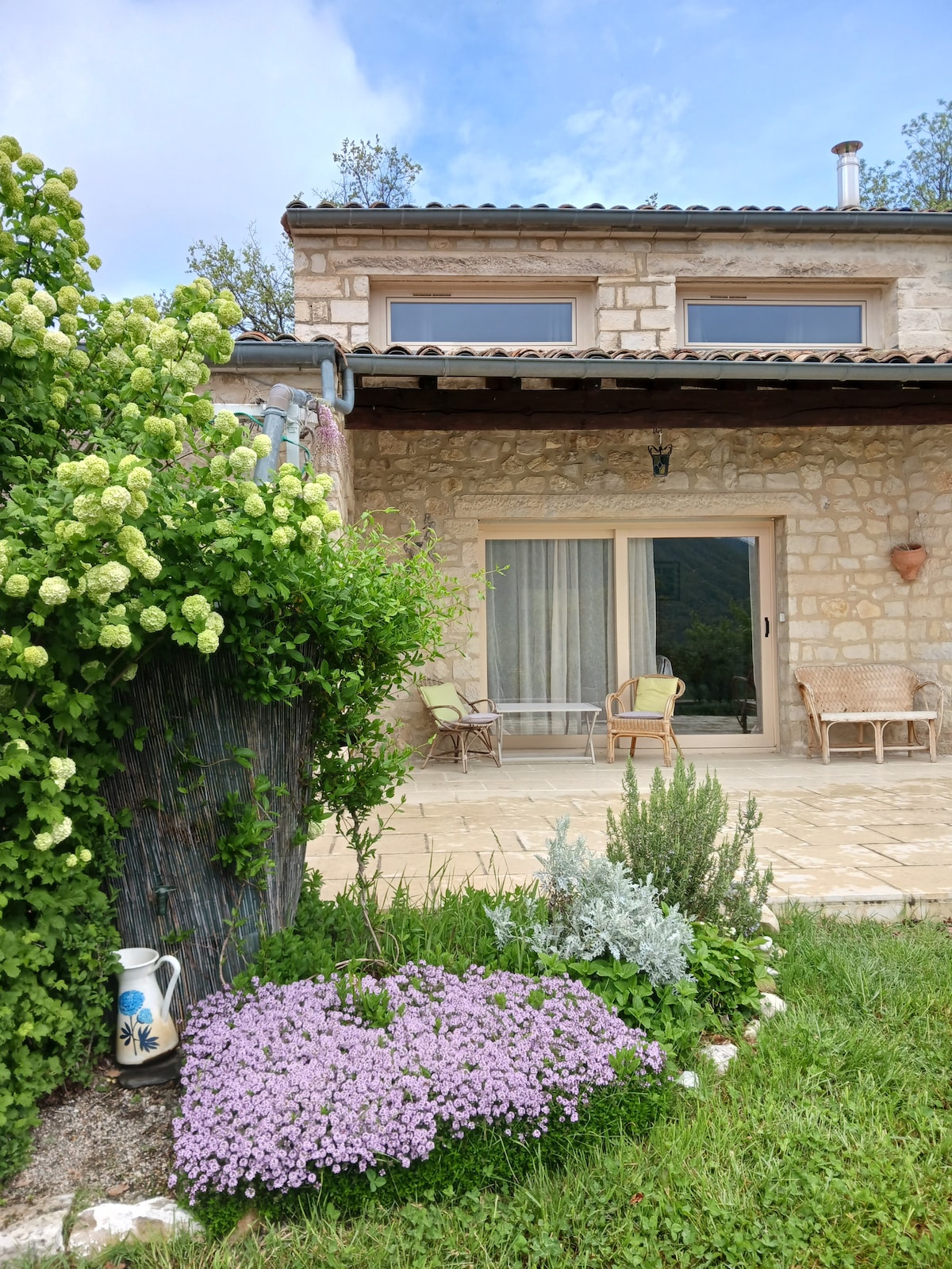 A stone house is framed by vibrant greenery and flowers. A spacious terrace features comfortable seating areas. Flowering plants in shades of purple and green enhance the outdoor space. Large glass doors allow natural light to flood into the living area, offering views of the surrounding landscape.