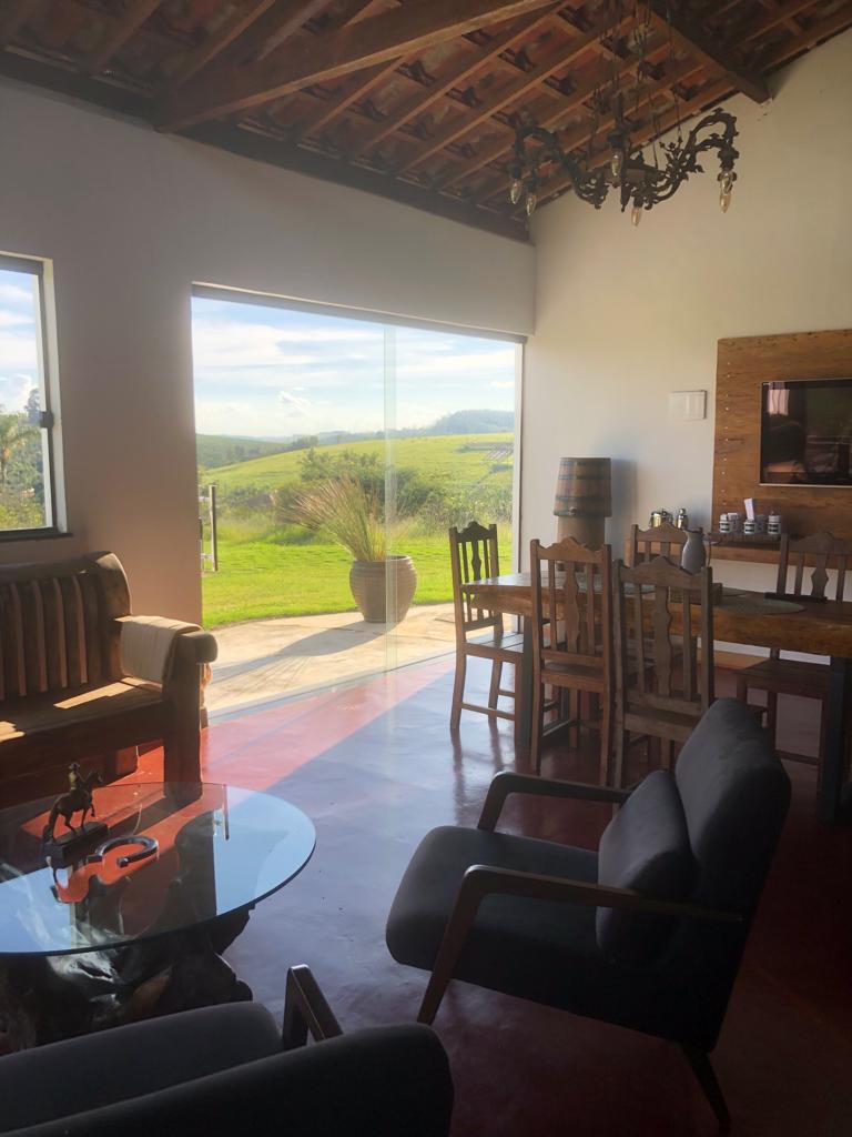 A bright living space features large windows that offer views of rolling hills and greenery. Wooden furniture, including a dining table and chairs, complements the rustic decor. A comfortable seating area is arranged around a glass coffee table, creating an inviting atmosphere.
