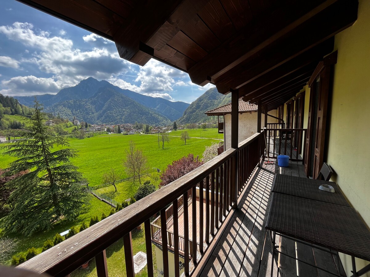 A balcony is featured, showcasing panoramic views of rolling hills and green fields. Wooden railings and beams frame the space, while sunlight casts shadows along the floor. Lush trees and distant mountains create a serene backdrop.