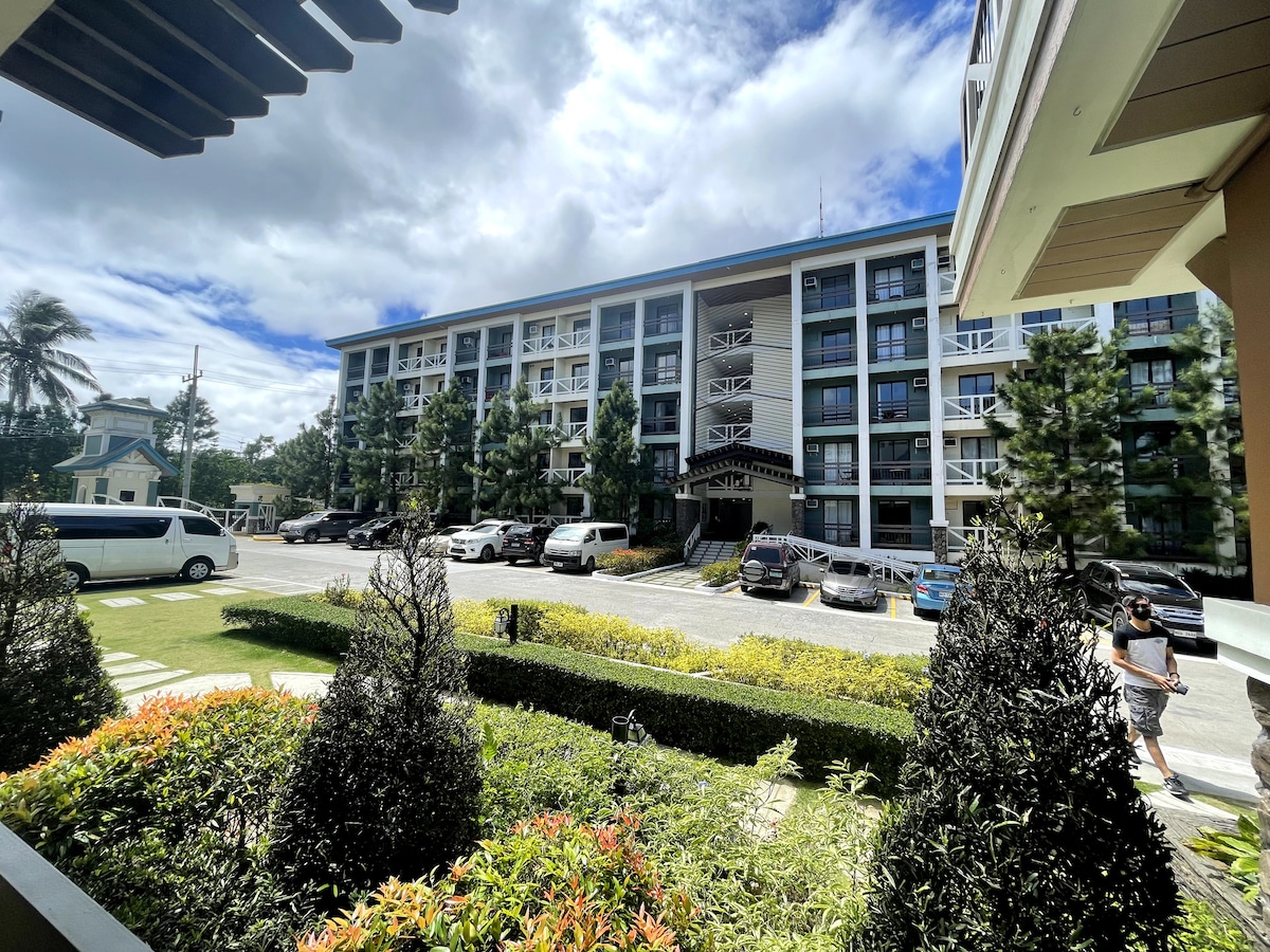 The exterior of the Pine Suites Tagaytay is visible, showcasing a four-story building surrounded by lush greenery and colorful landscaping. Multiple parked vehicles are seen in the parking area. A pathway leads through manicured shrubs, connecting to the entrance of the building.