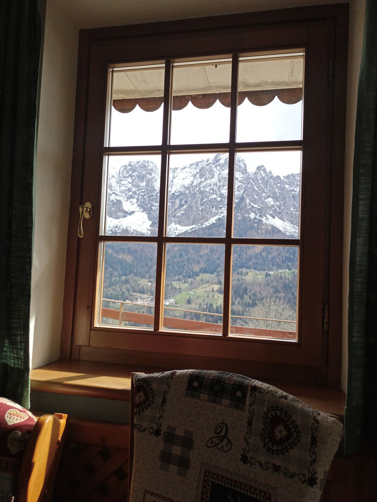 A wooden window frame showcases a stunning view of snow-capped mountains, surrounded by lush green valleys. Sunlight illuminates the scene, enhancing the natural beauty. Traditional curtains in deep green add a touch of coziness to the interior space.