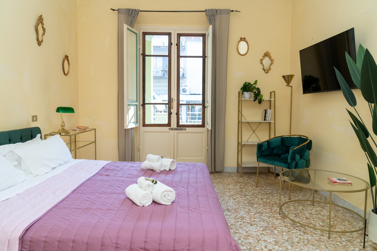 A well-appointed room features a spacious bed with a light pink quilt and neatly arranged towels. A cozy armchair is positioned beside a round glass table. A wall-mounted smart TV offers modern entertainment options, while large windows allow natural light to illuminate the space.
