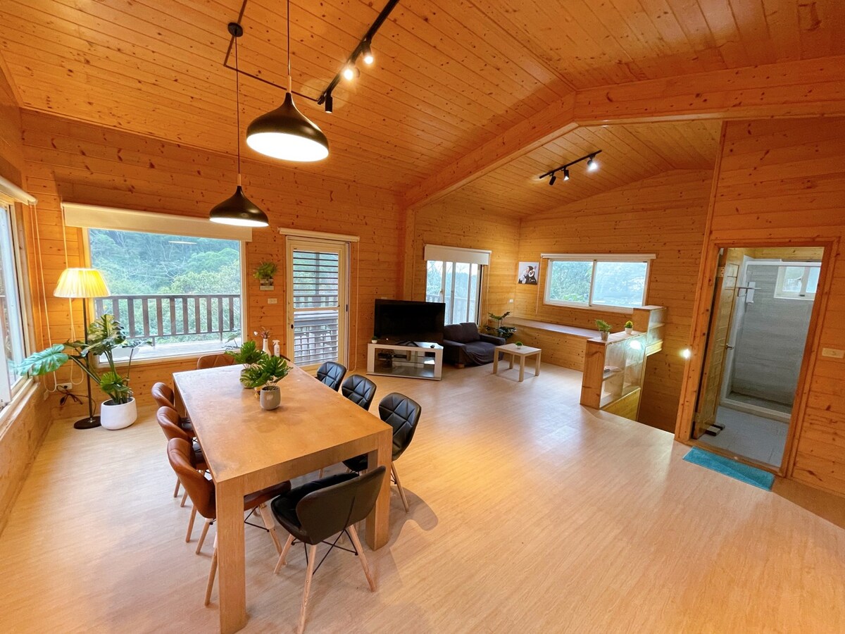 An open living area features natural wood paneling and high ceilings. A large dining table is surrounded by black chairs, while a comfortable seating area is positioned nearby. Windows bring natural light and views of the surrounding landscape. A television is visible along with potted plants.