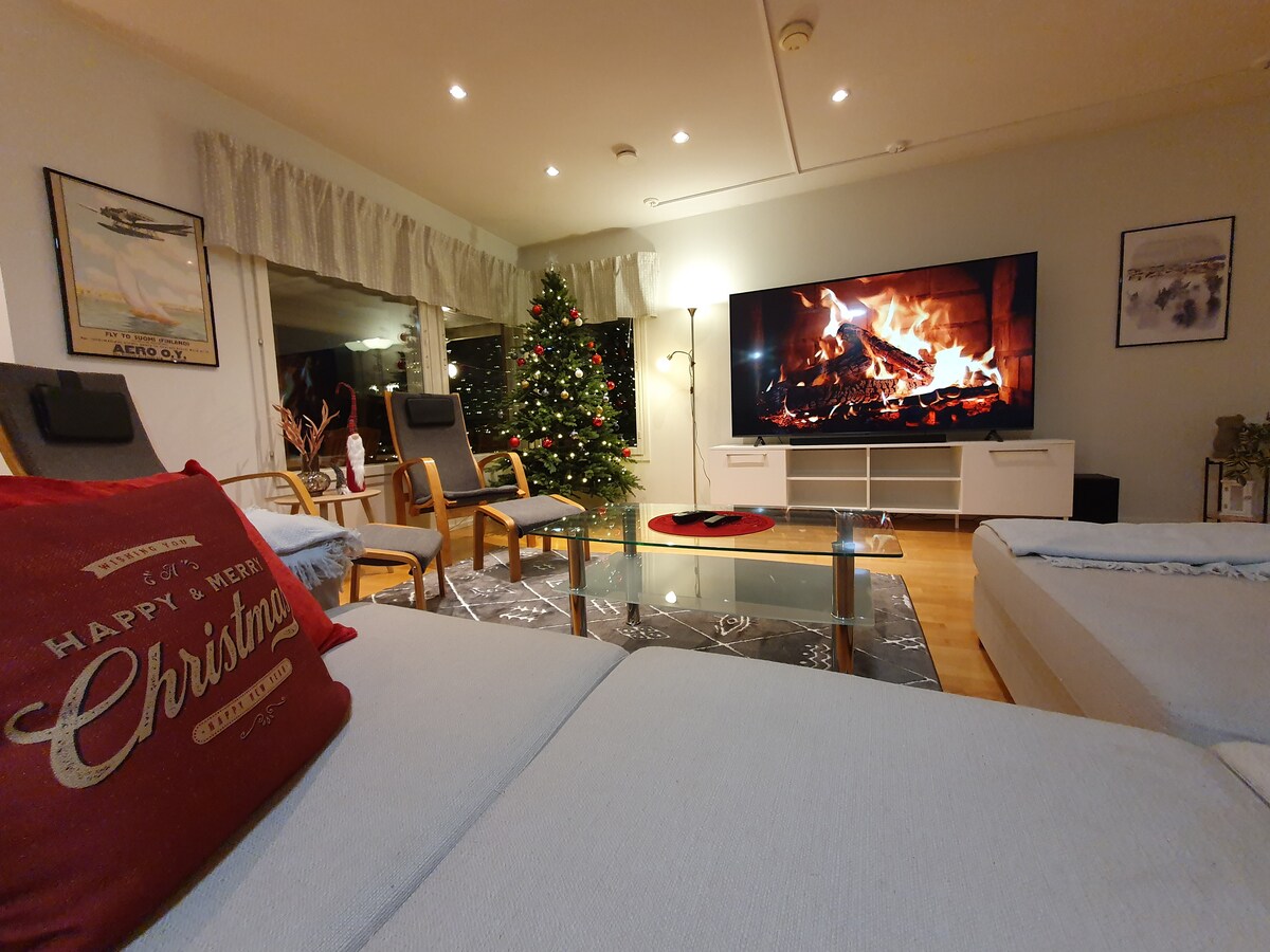 A cozy living area features a large television displaying a fireplace, creating a warm ambiance. Holiday decorations include a Christmas tree, while comfortable seating arrangements are positioned around a glass coffee table. Gentle lighting enhances the inviting space.