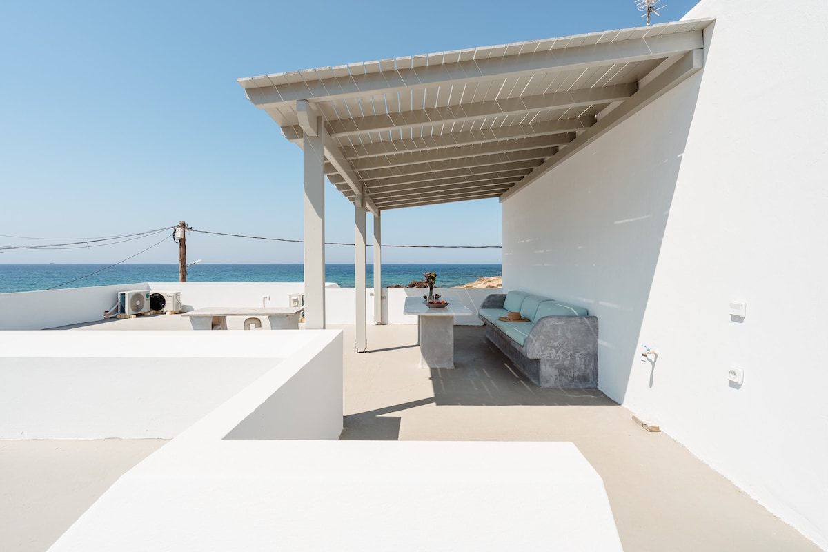 An outdoor terrace is depicted with a whitewashed structure and a covered seating area. Soft blue cushions are arranged on a stone bench, and a small table holds a potted plant. Clear views of the sea and sky extend beyond the terrace.