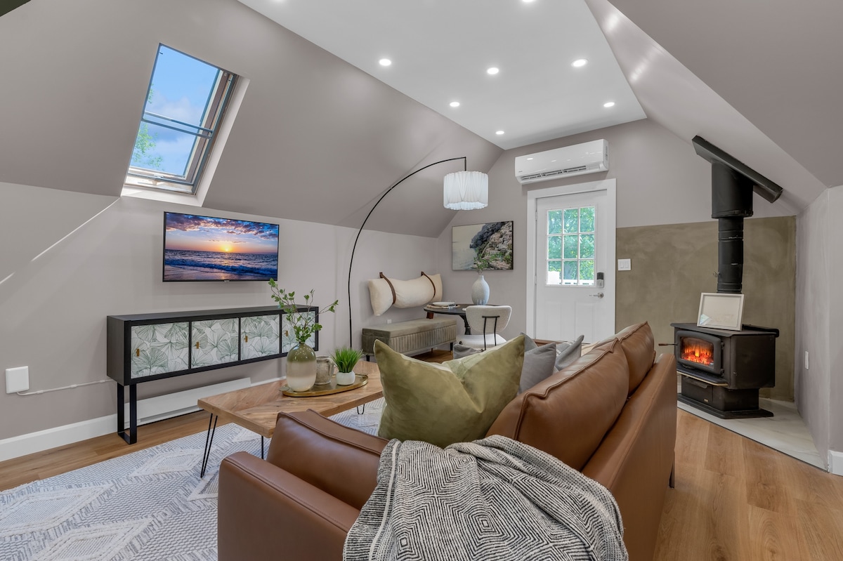 A cozy living area features a brown leather sofa complemented by soft cushions and a textured throw. Natural light floods in through a skylight and windows, highlighting the modern decor, including a corner television and a wood stove. A coffee table sits atop a woven rug.