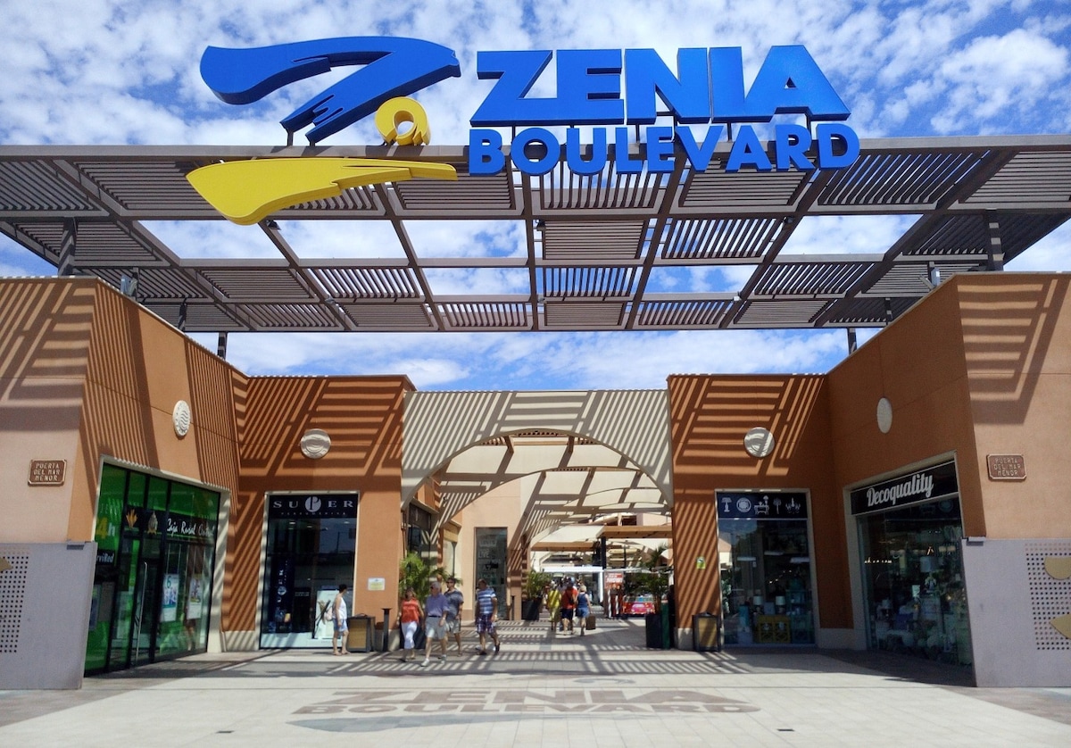 An entrance to Zenia Boulevard is shown, featuring a large blue sign overhead. The open walkways are lined with storefronts and greenery, creating a welcoming shopping environment. Bright blue skies and scattered clouds provide a pleasant backdrop.
