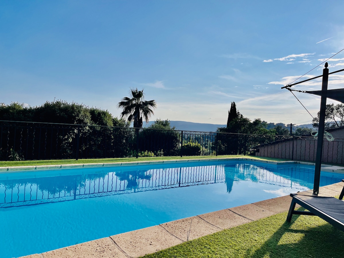 A serene swimming pool is featured, surrounded by a well-maintained green lawn. Palm trees and lush vegetation are visible in the background, framing a peaceful view of the landscape. The clear blue sky reflects on the water's surface, creating a tranquil setting.