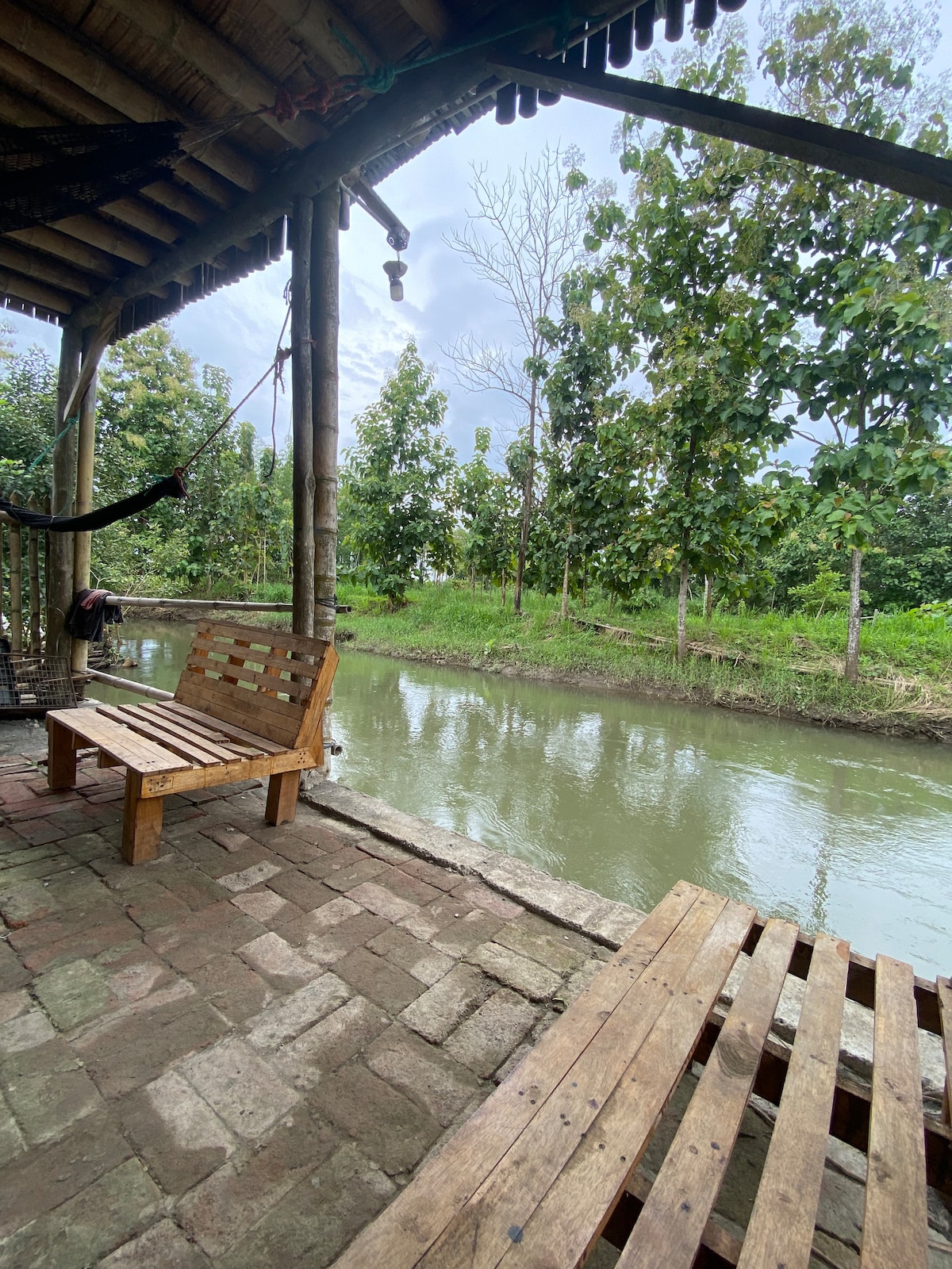 Two wooden benches are positioned along the edge of a gentle riverbank, offering a peaceful place to sit. Lush green trees line the water's edge, with a tranquil river reflecting the overcast sky. Cobblestone flooring complements the natural setting.