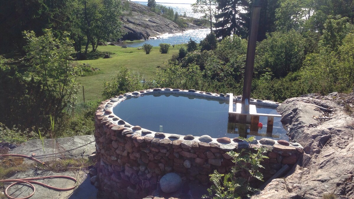 A natural stone hot tub is set on a rocky surface, surrounded by lush greenery. The still water reflects the sky, with glimpses of the ocean visible in the distance, creating a serene outdoor space for relaxation.