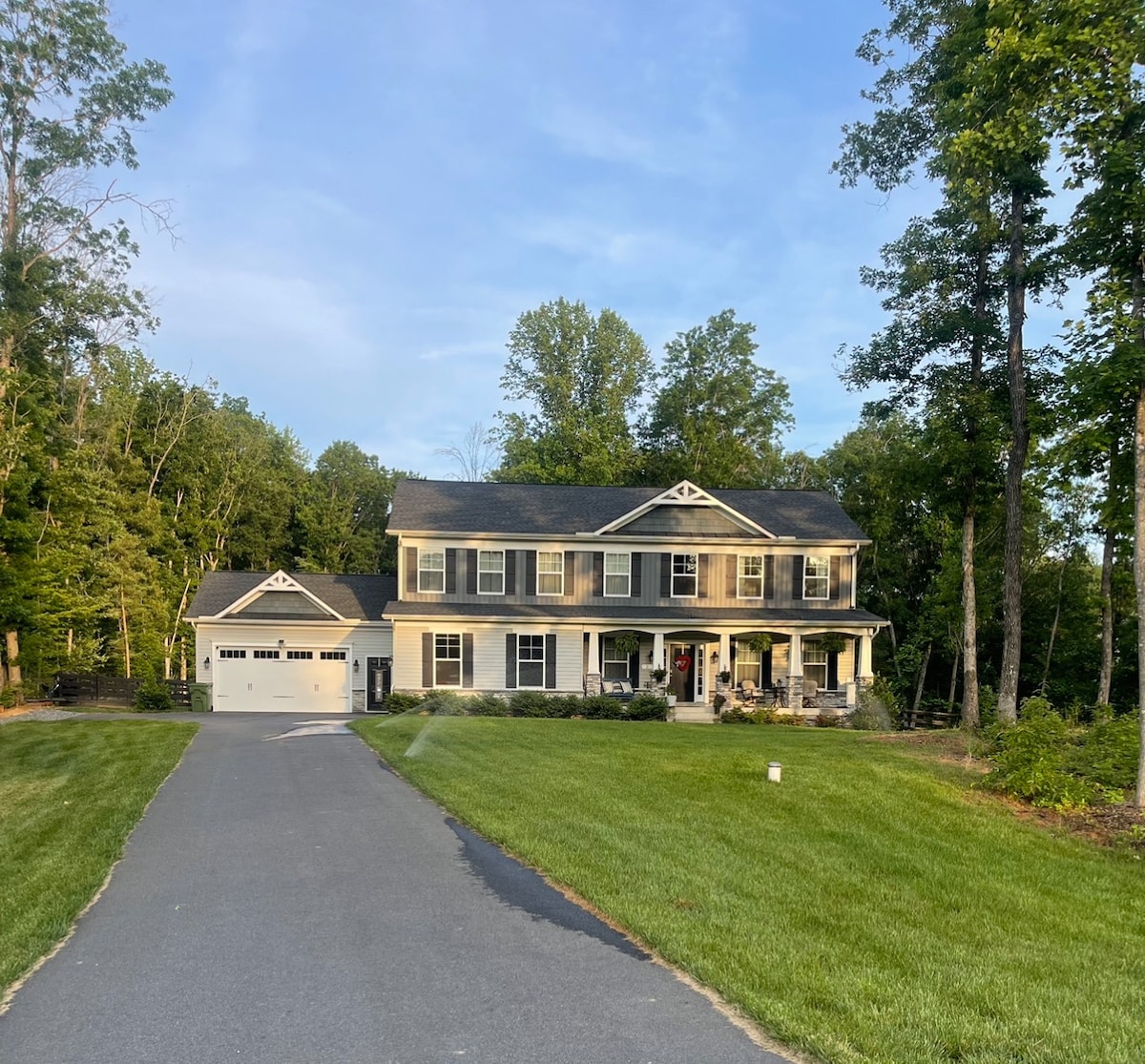 A modern two-story house is showcased, surrounded by lush greenery. A wide driveway leads to the home, which features a welcoming porch and rows of windows. The garage is visible on the left side, enhancing the overall spaciousness of the property.