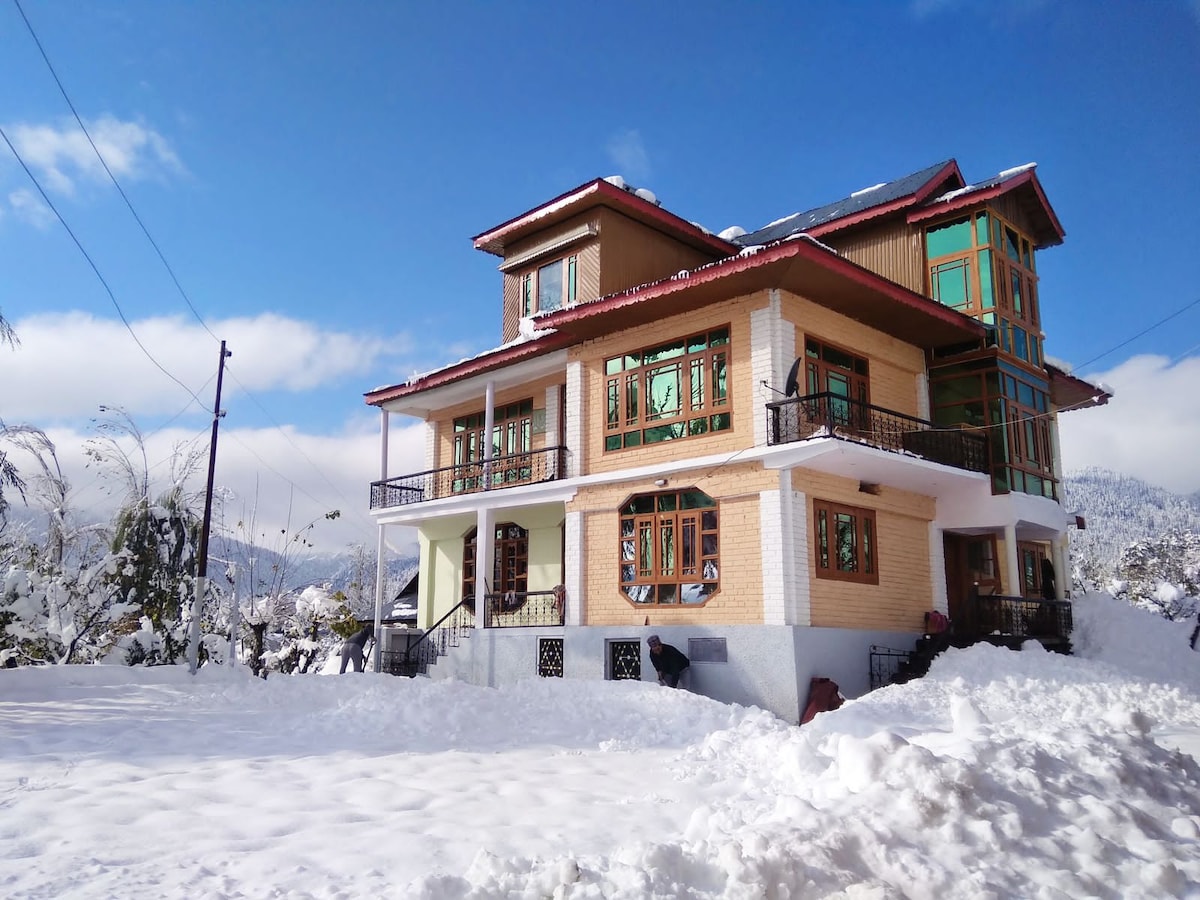 A spacious multi-level home is set against a backdrop of snow-covered mountains. Large windows framed in wood allow ample natural light, and balconies provide outdoor views. The structure's combination of wood and stone elements adds character to the calm winter landscape.
