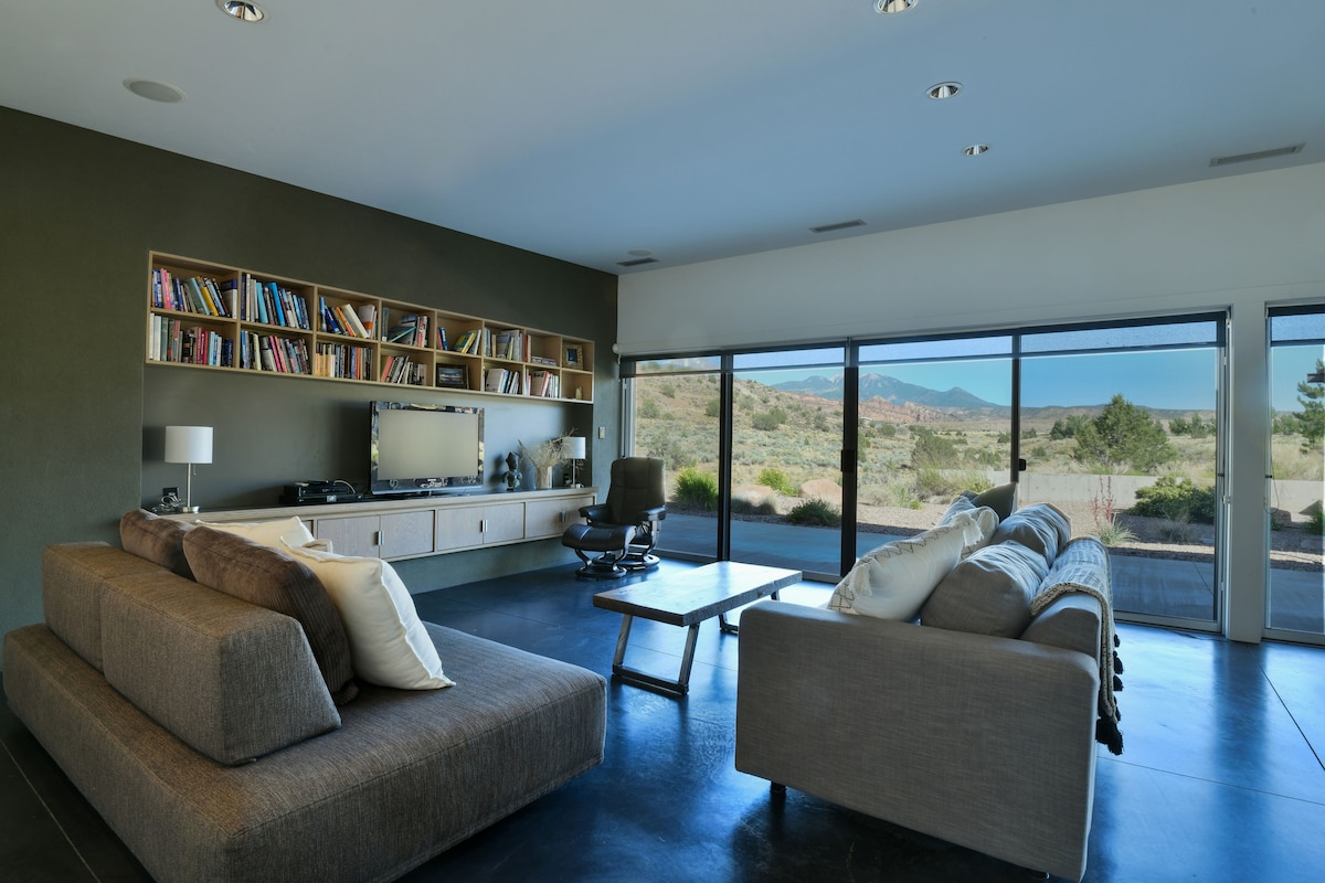 A modern living room features a comfortable sectional sofa and a coffee table, complemented by a media console and an office chair. Large windows provide natural light and stunning views of the surrounding landscape, ensuring a connection with the exterior environment.