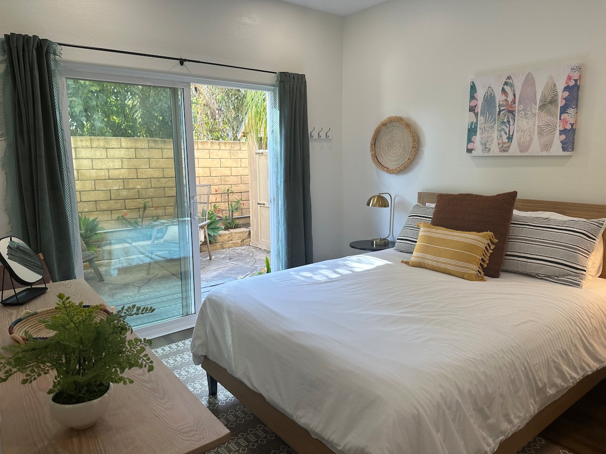A tranquil bedroom is adorned with a comfortable bed featuring neutral linens and decorative pillows. A sliding glass door opens to a private patio, enhancing the serene ambiance. Natural light filters through the curtains, and a small desk with a mirror is positioned nearby.