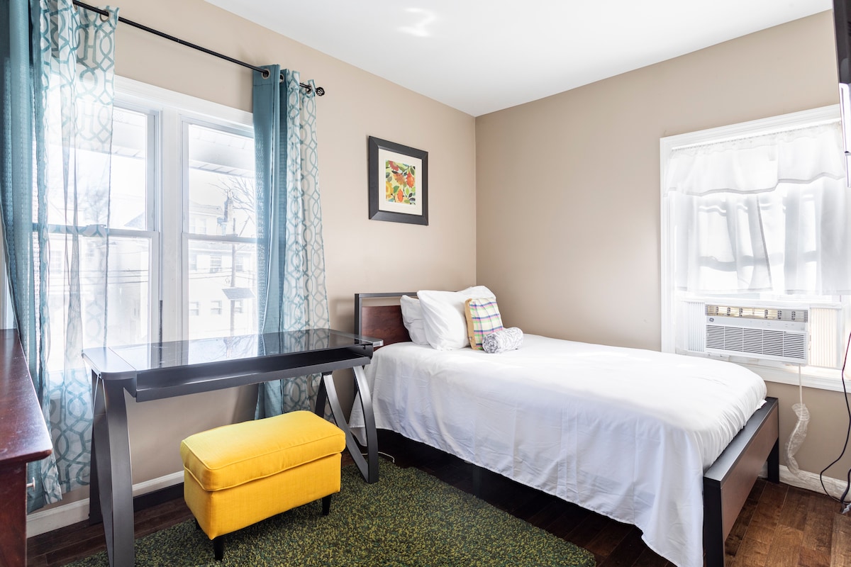 A bright sleeping area features a spacious bed with crisp white bedding, complemented by a yellow ottoman and a desk. Natural light filters through two windows with sheer curtains and teal-patterned drapes, illuminating the neutral wall color.