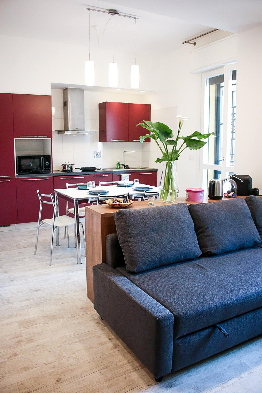 A modern kitchen area features red cabinetry, stainless steel appliances, and a dining table setup for four. A plush sofa in dark fabric is positioned in the foreground, with a vase of greenery placed nearby, complementing the warm ambiance of the space.