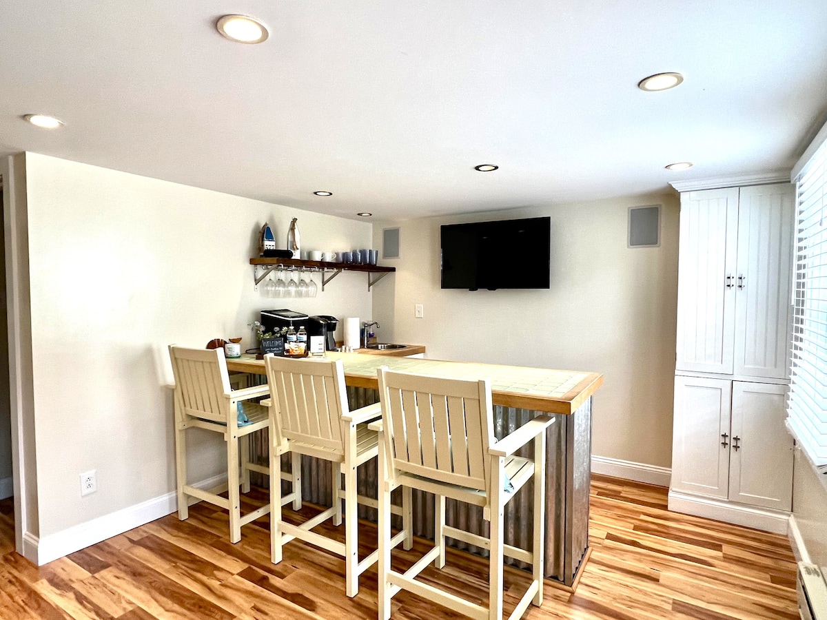 A clean, open kitchen and living area features a large bar with four white wooden stools. Shelving above holds glassware, and a television is mounted on the wall. Light wood flooring reflects natural light from the windows along one side of the room.