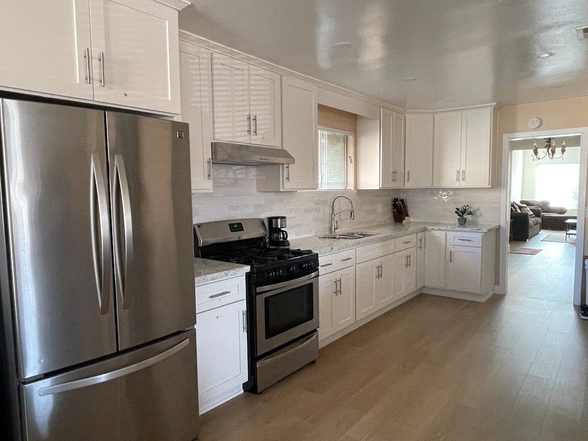 The kitchen features modern stainless steel appliances, including a refrigerator and stove. White cabinetry complements a marble-like countertop. A window allows natural light, and a coffee maker is visible on the counter. The open layout leads to the adjacent living space.