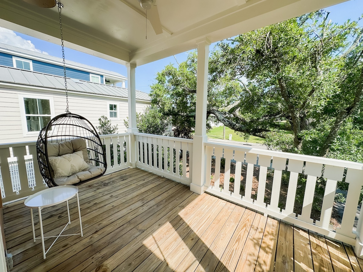 A spacious porch is presented with a wooden floor and white railings. A hanging chair is positioned in one corner, accompanied by a small side table. Lush greenery is visible through the railing, adding a serene outdoor view, with sunlight filtering into the space.