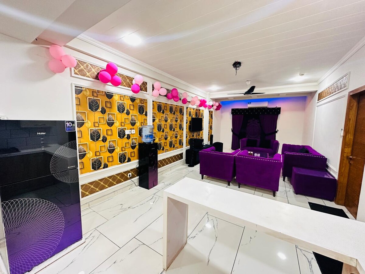 The living area features a vibrant design with purple furniture and playful pink decor. A modern entertainment center is positioned against an accent wall adorned with a bold pattern. Bright lighting highlights the spacious layout, complemented by sleek tile flooring.