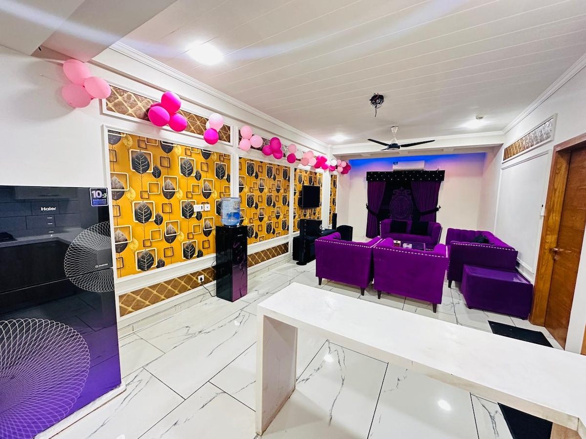 A vibrant living area showcases purple seating arrangements, complemented by a distinct yellow and black patterned wall. Decorative balloons hang above, and large windows allow natural light to fill the space. A sleek, white dining table contrasts with the colorful decor.