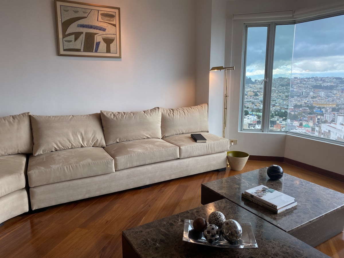 A spacious living area is furnished with a comfortable beige sectional sofa positioned to offer views through large windows. A modern coffee table with decorative items is centered in the room, complemented by a floor lamp that adds soft lighting. A piece of artwork is displayed on the wall.