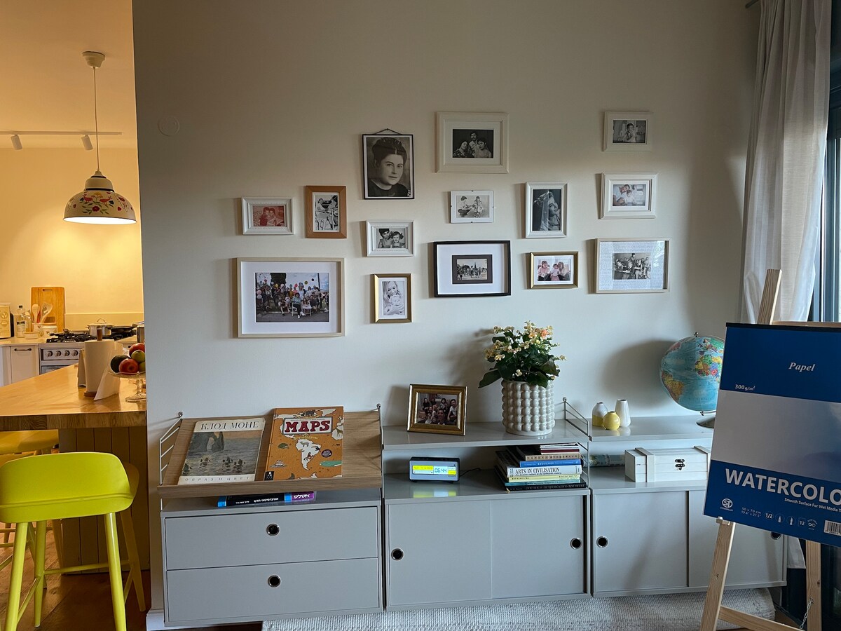 A neatly arranged wall displays a collection of framed photographs in various sizes. Below, a console is topped with books, a small flower arrangement, and a globe. A bright yellow stool and an art sign complete the scene, adding color to the room.