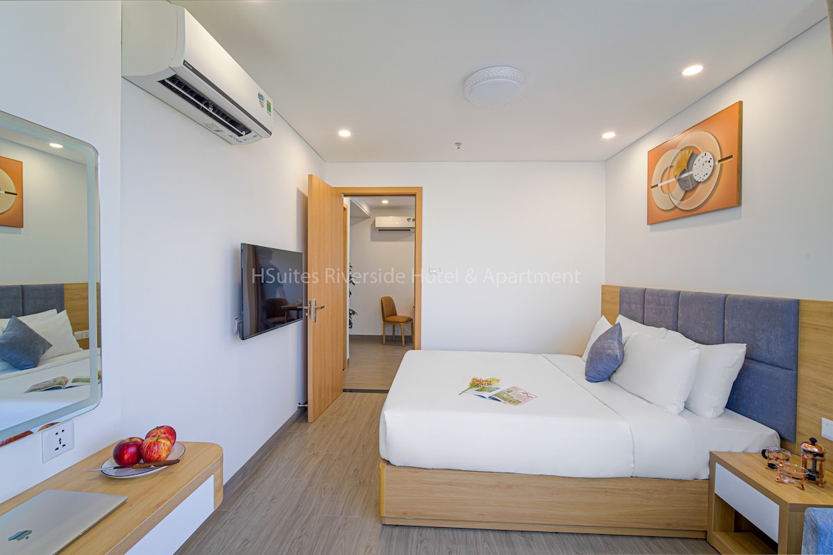 A bright and airy bedroom features a queen-sized bed with soft white linens. A wall-mounted TV is positioned across from a wooden dresser. The space is complemented by two decorative wall art pieces and a large mirror reflecting natural light from the window.