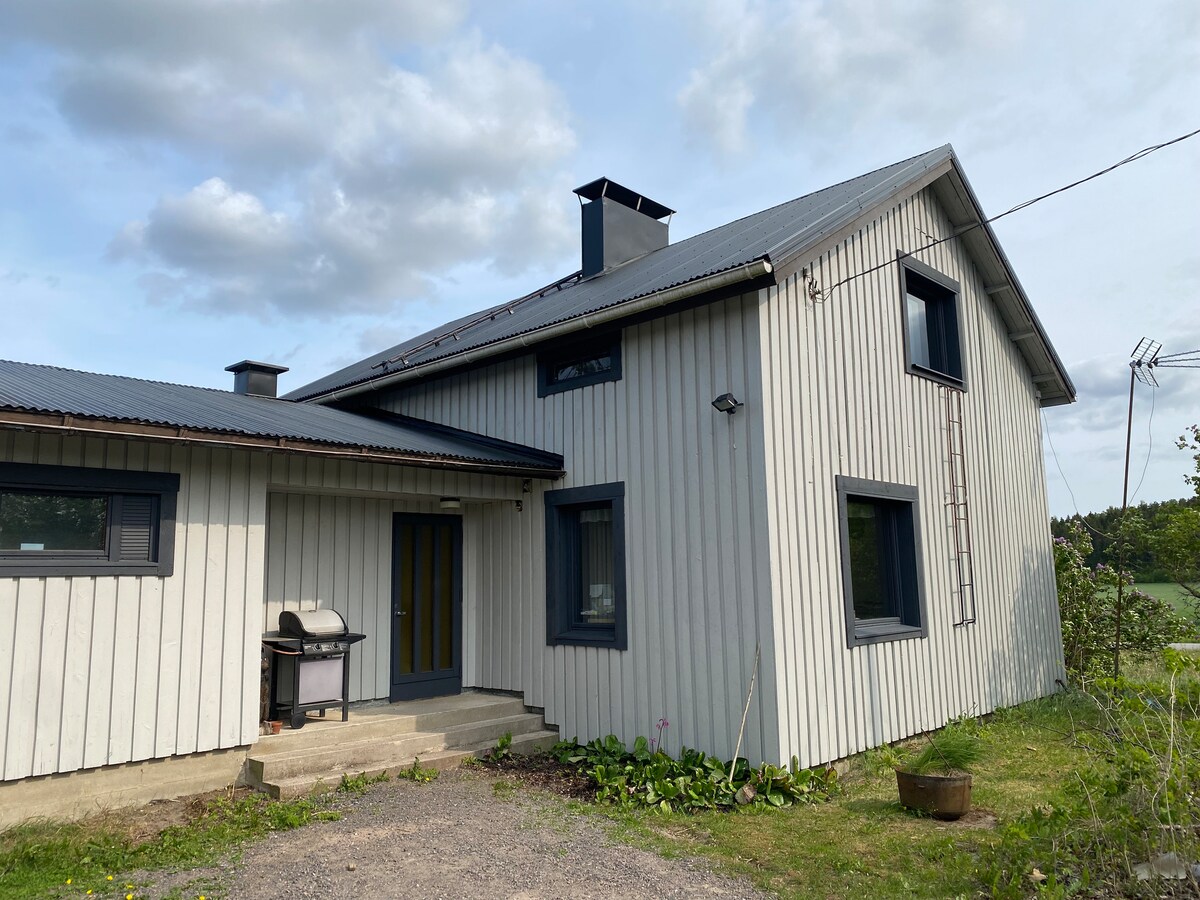 A modern two-story house is depicted, featuring a combination of grey wooden siding and large windows. A small outdoor patio area is visible, equipped with a grill. The surrounding landscape includes green areas and a clear sky with scattered clouds.
