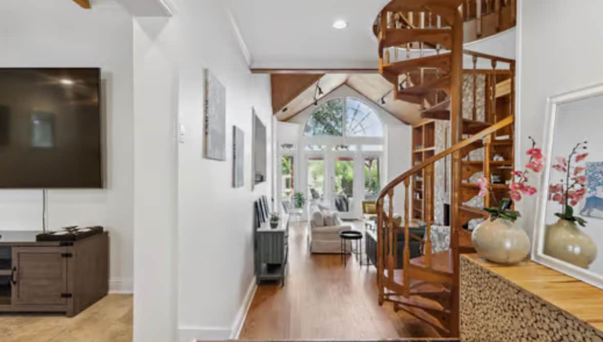 The entryway leads into a bright living area with large windows allowing natural light. A spiral staircase is prominently featured, connecting the two levels. Light wooden flooring provides a warm atmosphere, while contemporary art adorns the walls, enhancing the space's open feel.