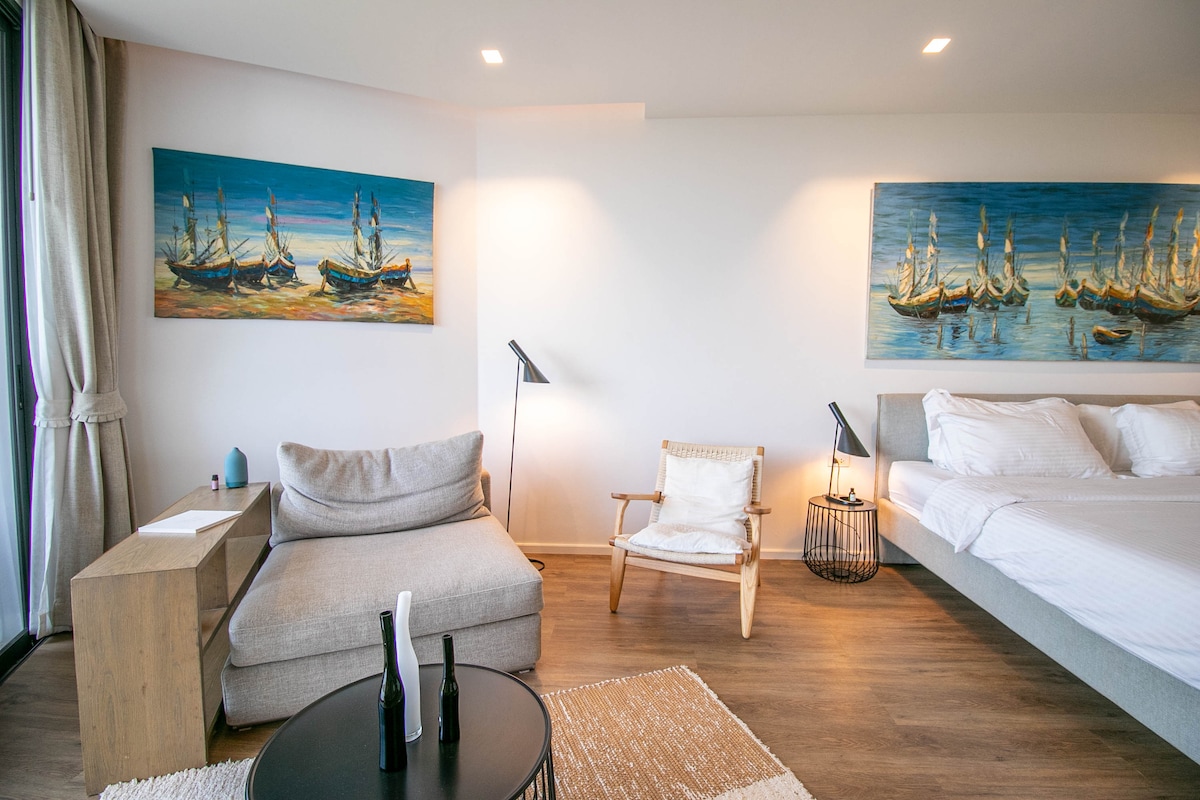 A spacious room features a comfortable seating area with a light-colored sofa and a simple wooden coffee table. Two artistic paintings of boats adorn the walls, complemented by a modern accent chair. Soft lighting highlights the cozy decor and inviting atmosphere.