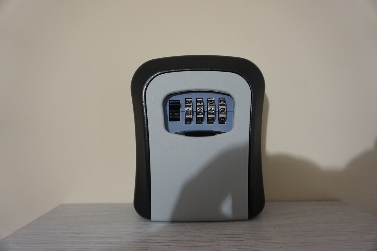 A secure combination lockbox is displayed against a plain backdrop. The lockbox features a numeric keypad with four dials for inputting a security code, designed to store valuables safely. It rests on a light-colored wooden surface.