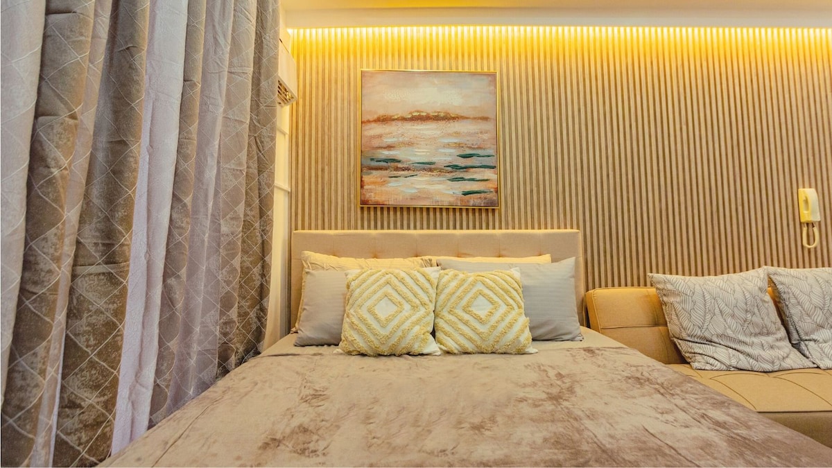 A modern sleeping area features a spacious bed adorned with soft, neutral bedding and decorative pillows. A cozy couch complements the space, while a piece of artwork adds a splash of color to the warm-toned wall with textured paneling.