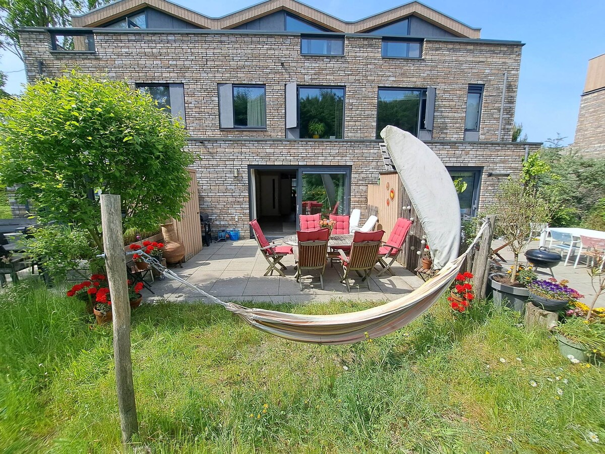 A spacious outdoor area features comfortable seating arranged around a stone patio, surrounded by vibrant flowers. A hammock hangs between wooden posts, offering a relaxing spot. The modern building showcases large windows, providing views of the green garden and inviting outdoor space.