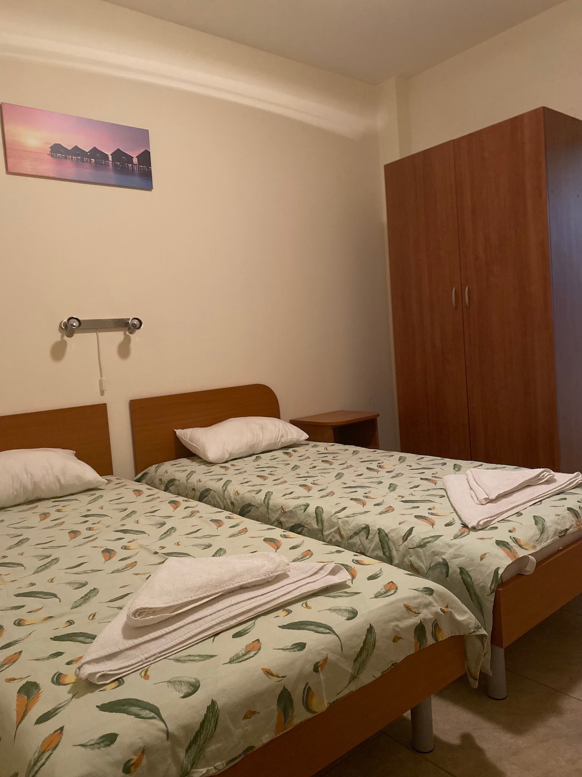 A sleeping area is equipped with two twin beds, both dressed in floral-patterned bedding. A large wardrobe is positioned nearby, providing ample storage space. The wall features a serene landscape image above the beds, while soft lighting enhances the overall ambience.
