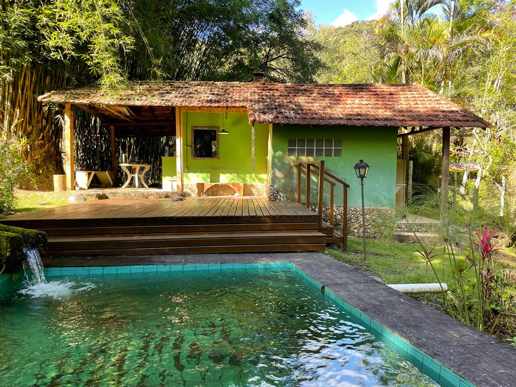 A rustic house with a green exterior and a porch is surrounded by lush vegetation. A wooden deck leads to a natural pool, with water gently cascading from a nearby feature. The tranquil setting is accentuated by the sound of nature.