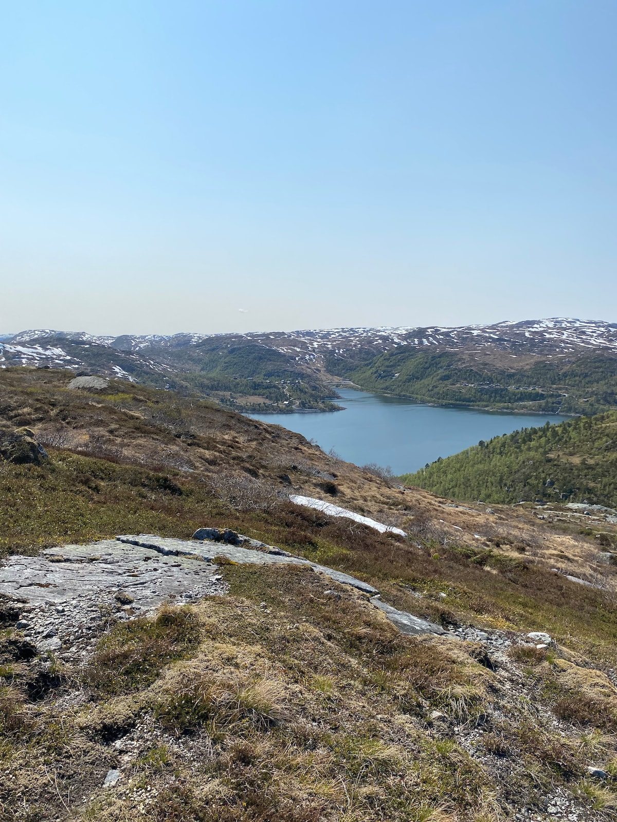 A scenic view features a serene lake surrounded by lush green valleys and distant mountains. Snow-capped peaks are visible in the background, while rocky terrain and scattered grasses dominate the foreground. The clear sky enhances the tranquil atmosphere of the natural landscape.