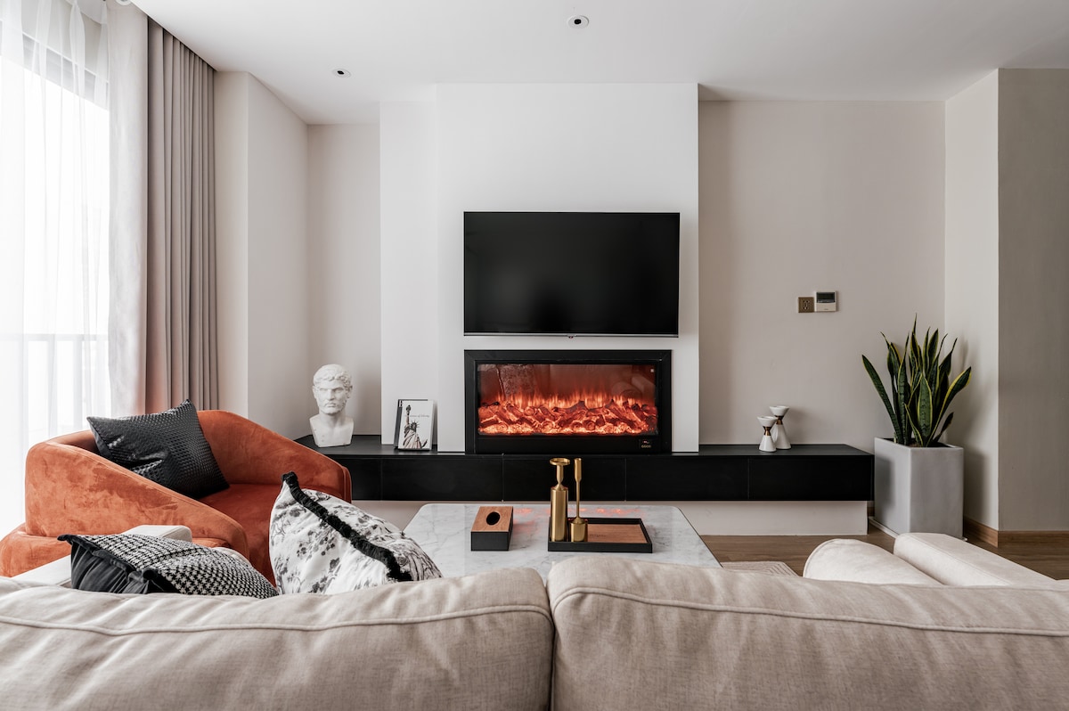 A cozy living area features a large, soft sofa facing a modern electric fireplace, which is complemented by a wall-mounted flat-screen TV. A potted plant adds a touch of greenery, while decorative items are thoughtfully arranged on a stylish coffee table.