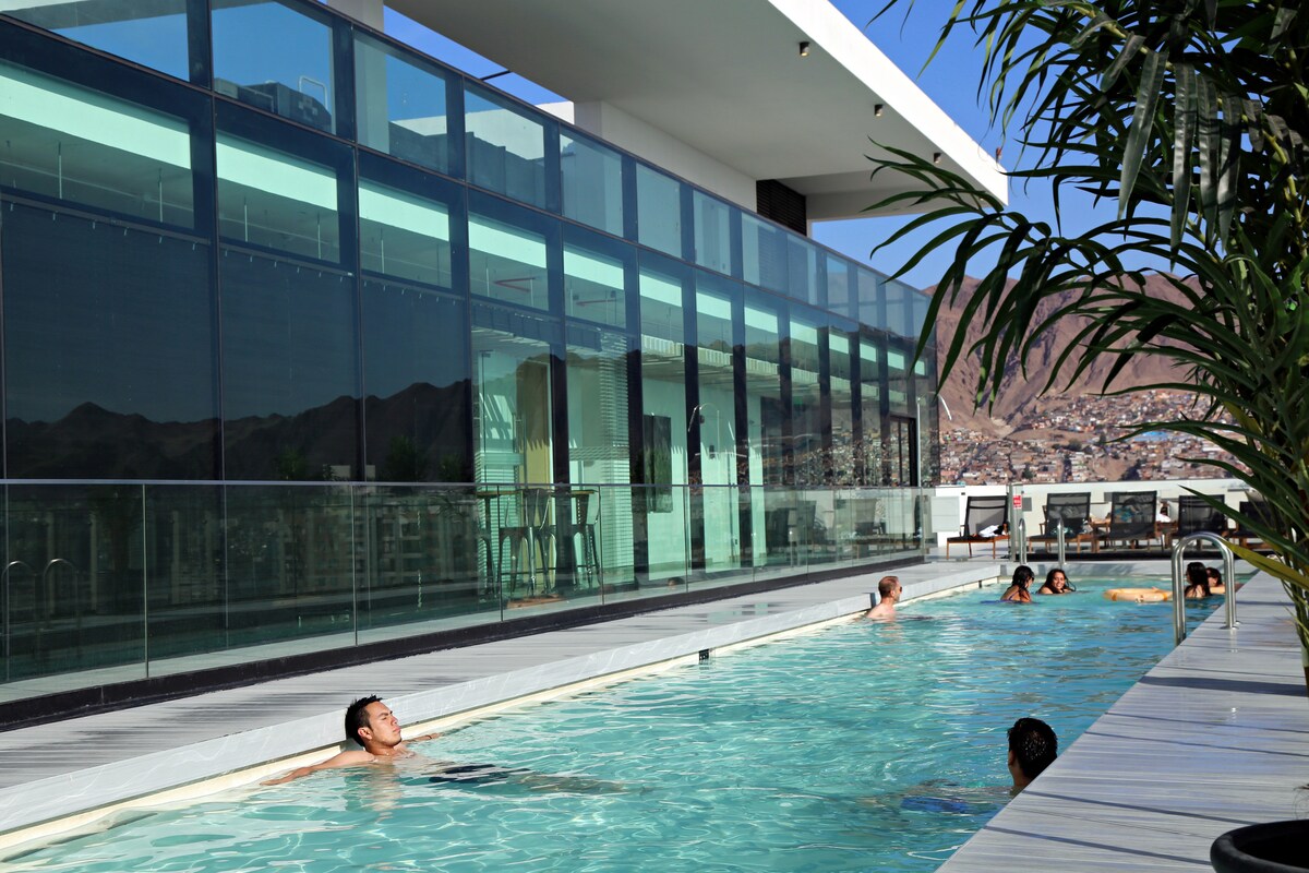 A modern rooftop pool area is visible, framed by sleek glass walls. Sunlight reflects off the calm water, which is surrounded by a spacious deck equipped with lounge chairs. Nearby, plants add a touch of greenery to the contemporary setting, while scenic views of the surrounding cityscape can be seen.