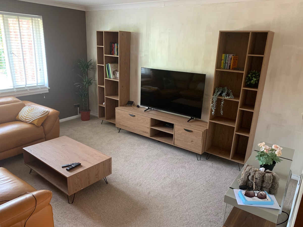 A comfortable living room is showcased, featuring a large flat-screen television mounted above a low wooden media unit. Two wooden bookshelves flank the TV, displaying books and decorative items. A cozy orange sofa and coffee table create a welcoming seating area, complemented by soft carpeting.