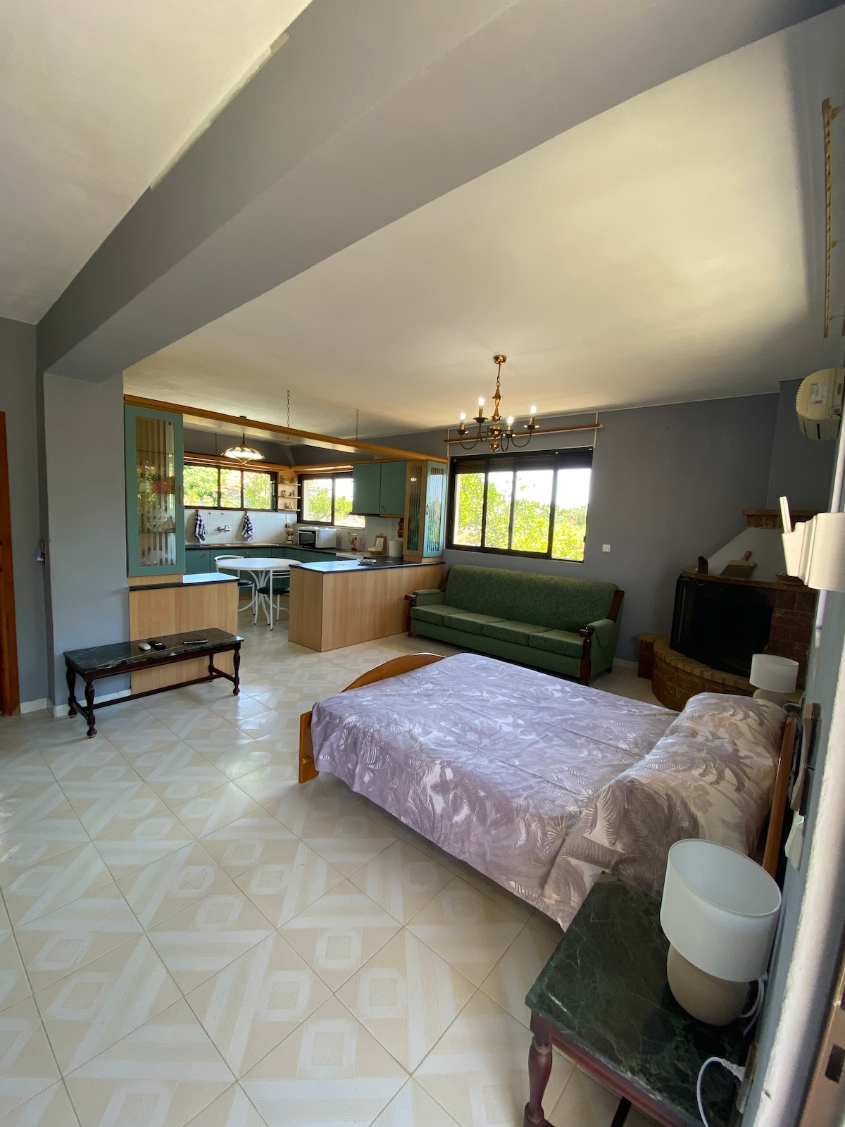 A spacious living area is featured, combining a cozy sleeping space with a soft comforter and an inviting green sofa. A well-equipped kitchen with modern appliances is visible in the background, complemented by a dining area with three stools. Natural light enters through large windows.