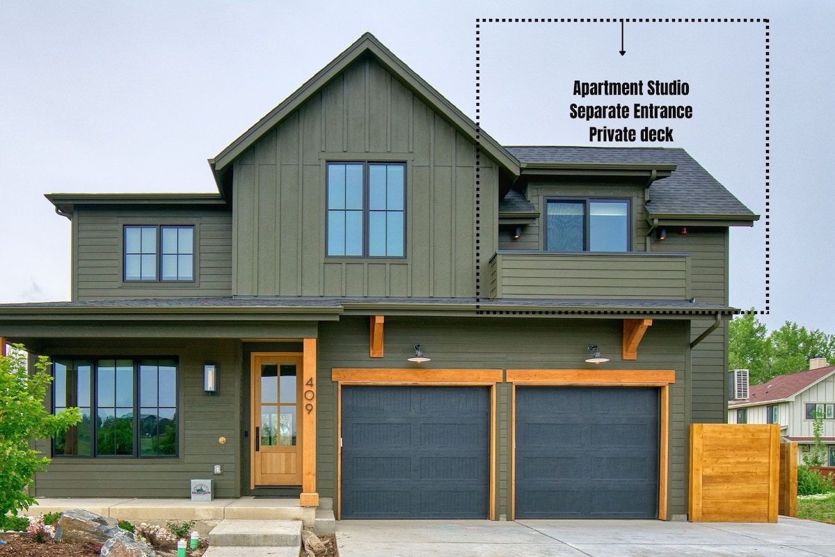 A modern two-story building features a separate entrance leading to the studio apartment. The exterior is clad in dark green siding, accentuated by wooden beams. A private deck is visible above the garage, enhancing the overall design of the residence.